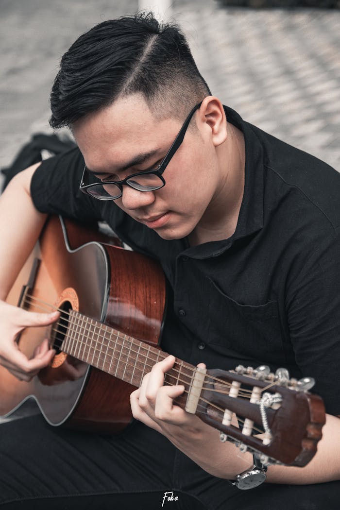 A young man sits outdoors playing an acoustic guitar, deeply focused and passionate.