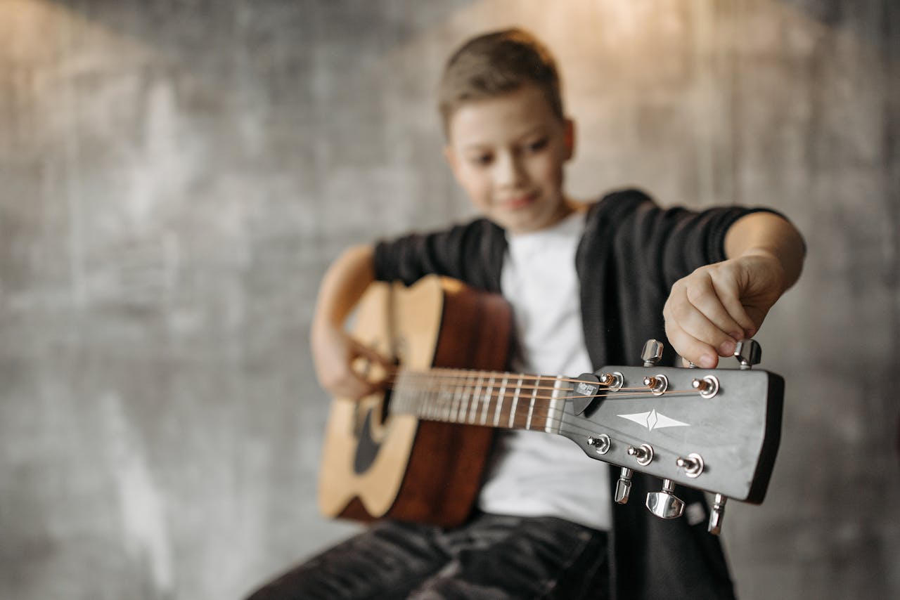 A young boy tuning an acoustic guitar indoors, focusing on musical learning.