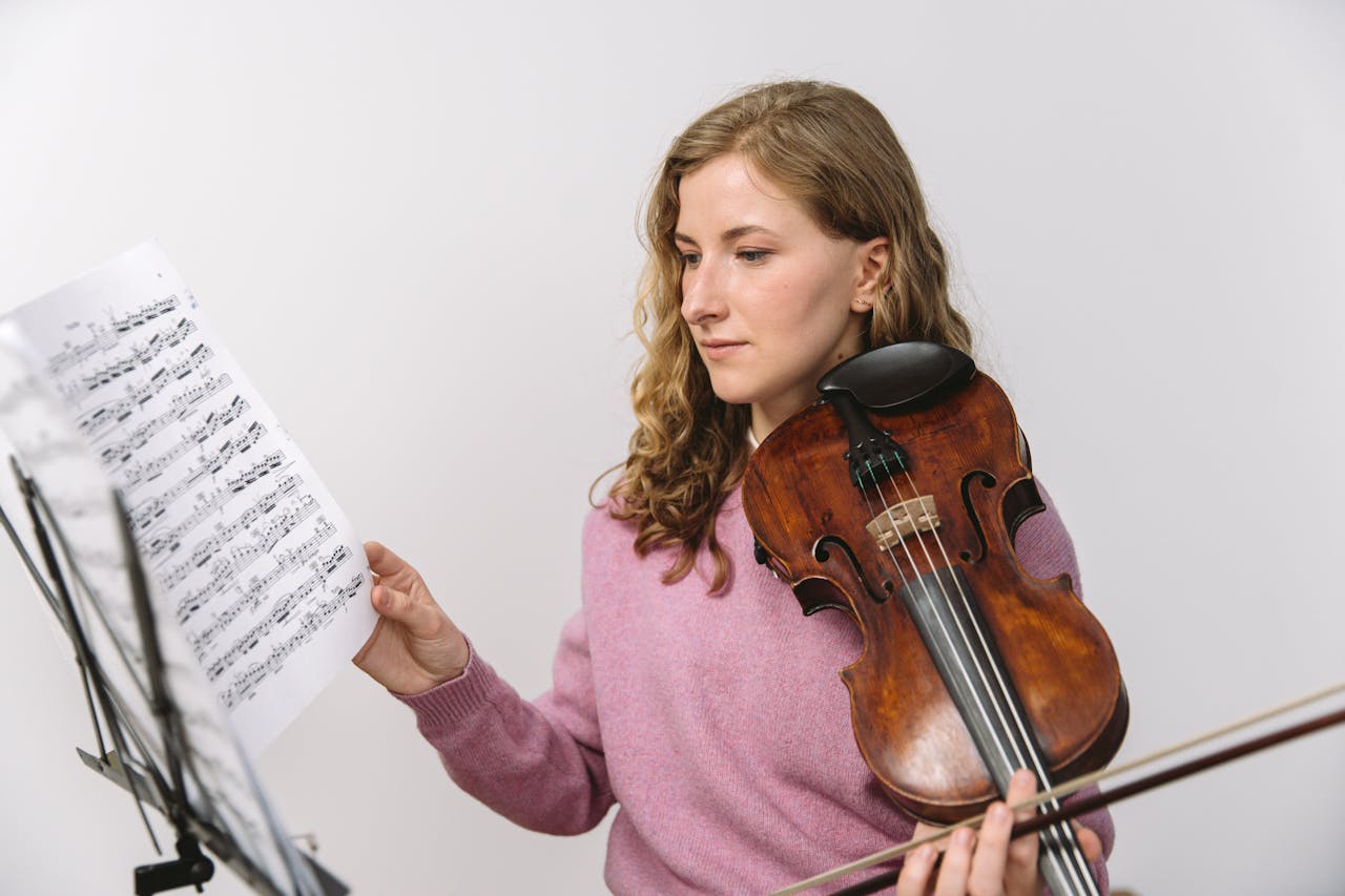 A female violinist in a pink sweater reads sheet music in a white studio, holding a violin.